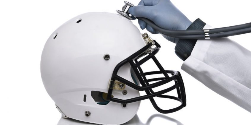 a football helmet and doctors hand holding a stethoscope on the crown of the helmet. sports concussion concept, and related conditions, cte, alzheimer's, parkinson's.